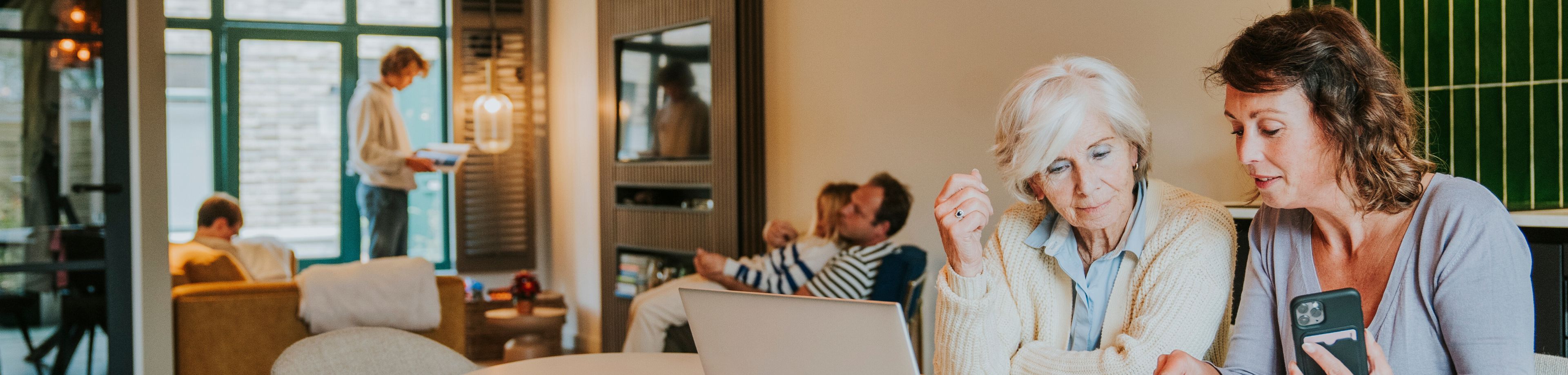 Een vrouw met telefoon in haar hand zit met haar moeder aan tafel achter een laptop.