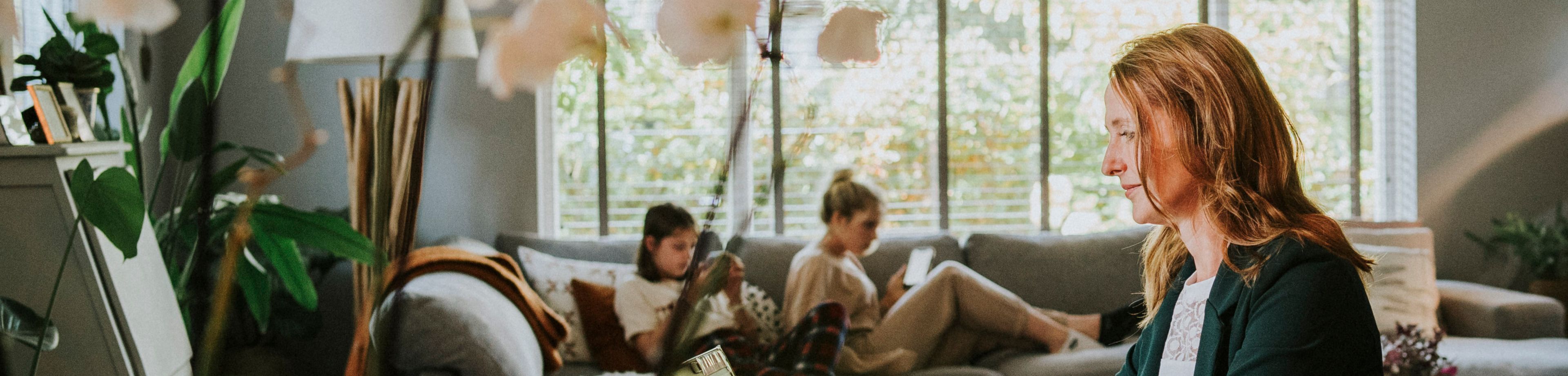 Een vrouw zit aan tafel en werkt op haar laptop. Op de achtergrond zitten 2 dochters op de bank, op hun telefoons te kijken.
