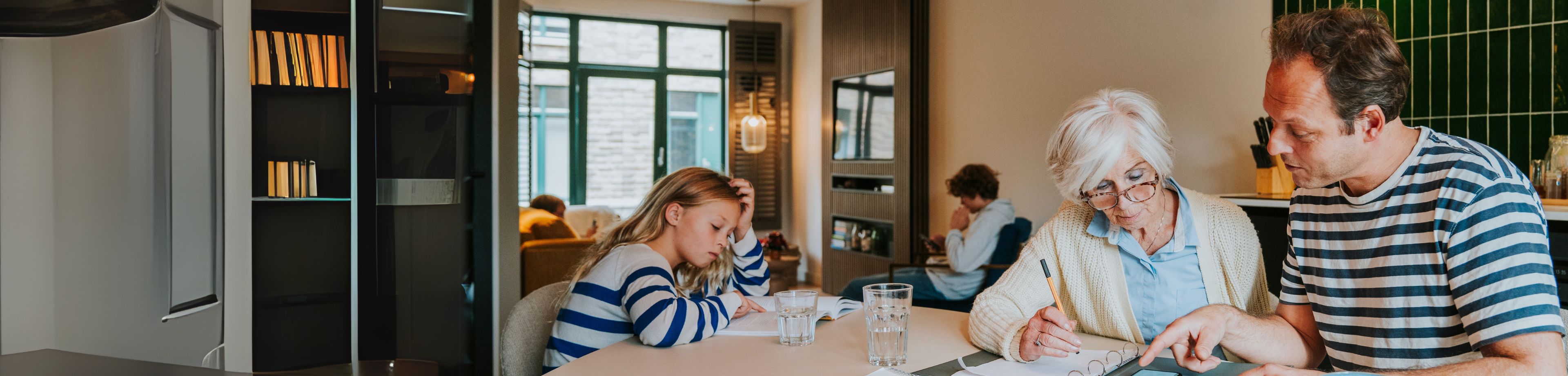Dochter leest een boek terwijl vader en oma samen documenten doornemen aan de keukentafel. 