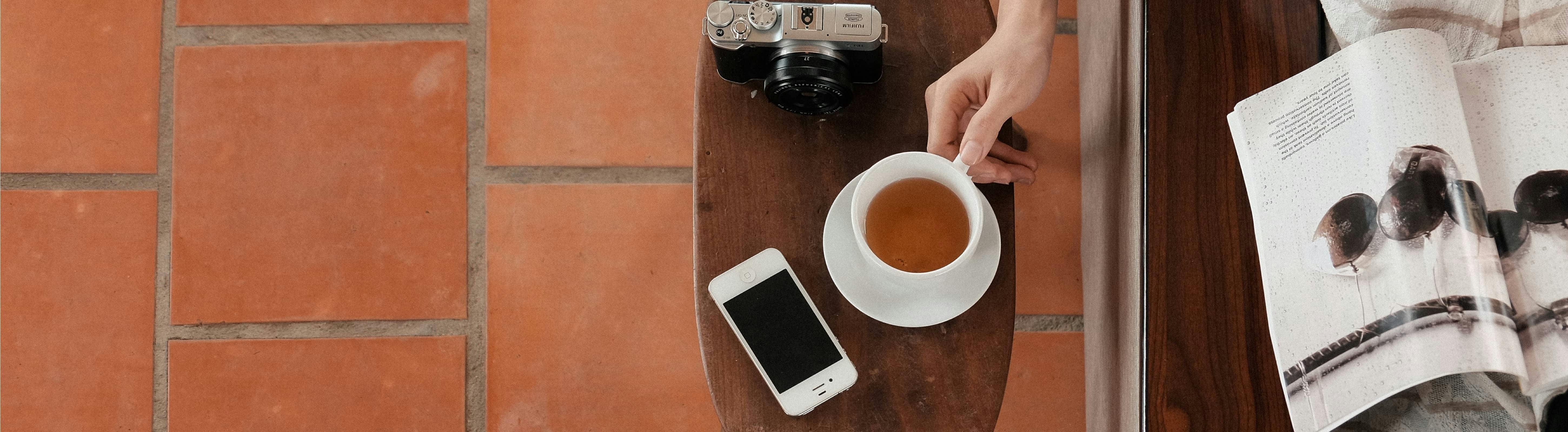 Vrouw zit aan een tafel een tijdschrift te lezen. Ze pakt haar kop koffie van de tafel waar ook een smartphone en een fototoestel liggen. Met de BuitenDeDeurverzekering® van Interpolis kunt u uw spullen buitenshuis in Nederland verzekeren.