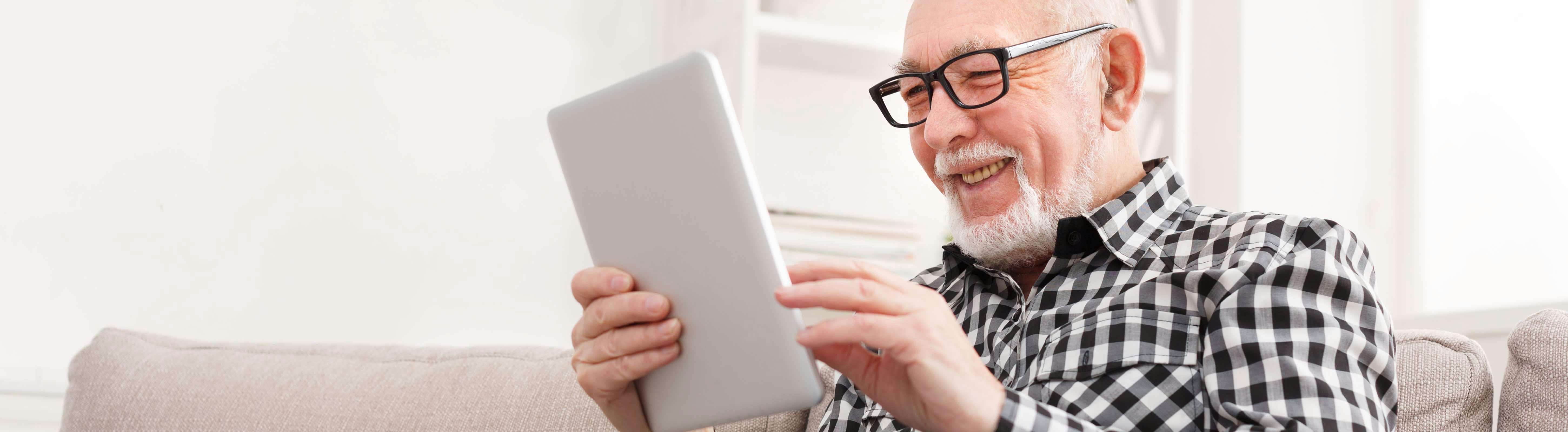 Man zit lachend op de bank met een tablet in zijn hand. Toekomstbestendig wonen, nu en later.