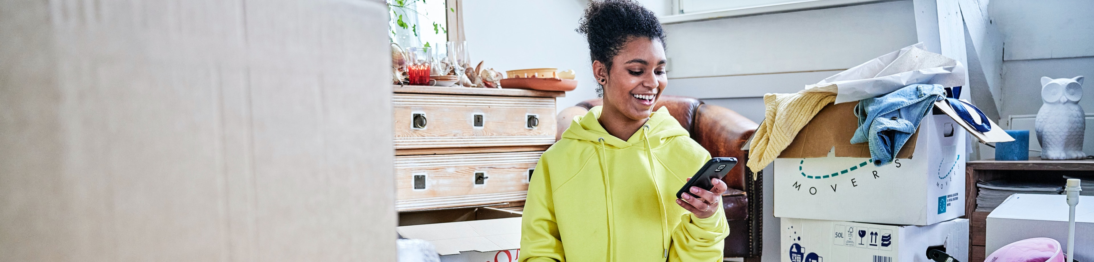 vrouw kijkt zittend tussen ingepakte verhuisdozen op haar smartphone
