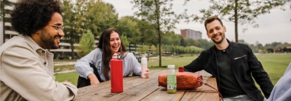 Vrienden zitten aan een picknicktafel in het park. 1 op de 4 millennials heeft burn-outklachten. Daarom onderzoeken we hoe we ervoor kunnen zorgen dat het aantal millennials met burn-outklachten afneemt.