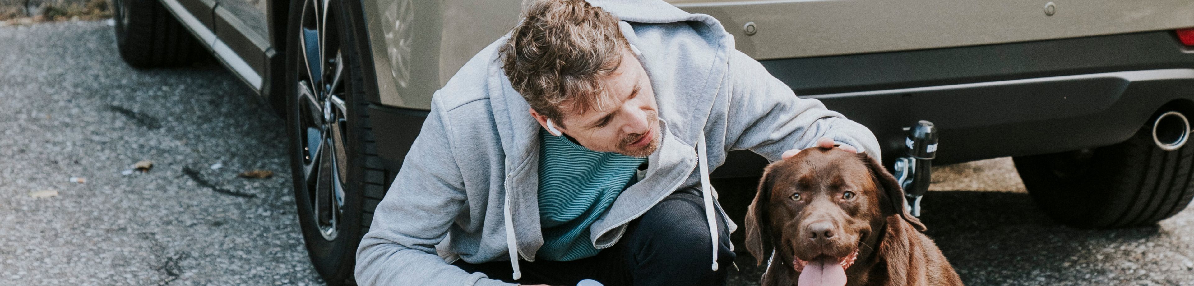 Een man aait een hond, vlakbij een geparkeerde auto met de koffer open