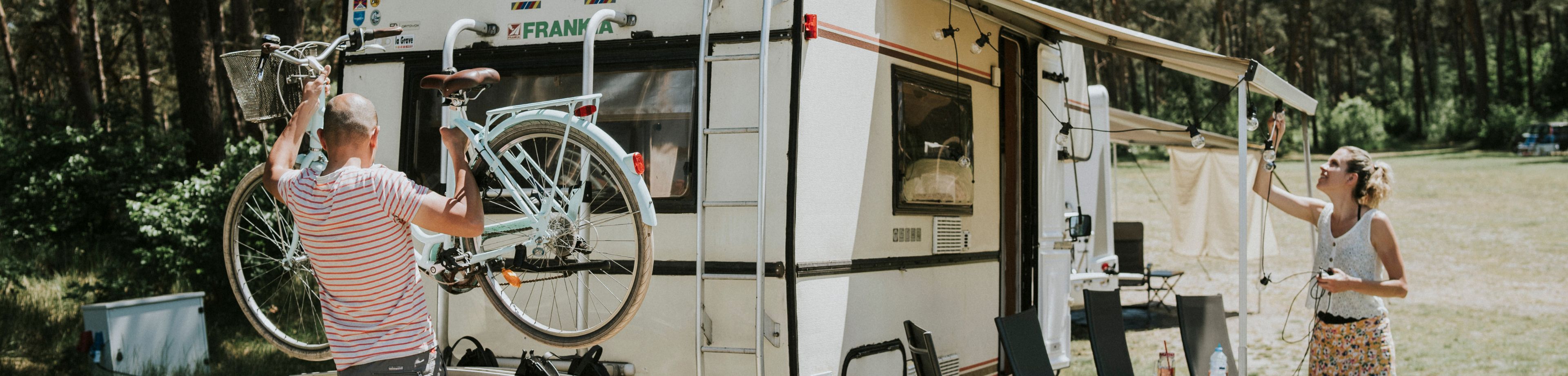 Man staat met fiets in zijn handen achter bij de fietsendrager van de camper en vrouw staat bij luifel met lampjessnoer