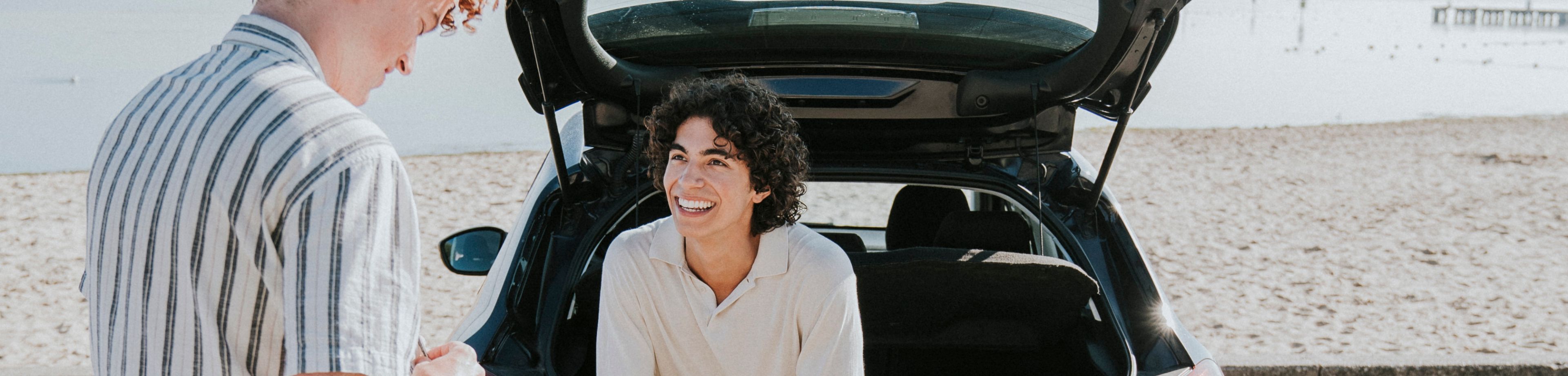 Jongen zit in de kofferbak van een auto dichtbij het strand staat geparkeerd. Een andere jongen staat ernaast. Samen lachen ze.