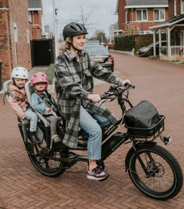 Een jonge vrouw zit op de fiets, met 2 kleine kinderen achterop. Ze hebben aandacht voor hun veiligheid, daarom dragen ze een fietshelm.