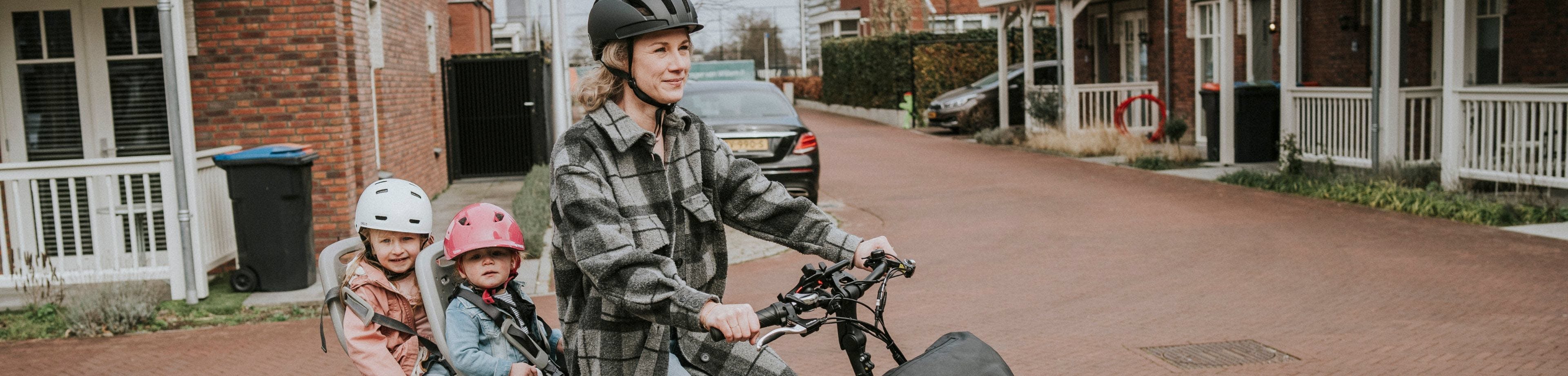 Een jonge vrouw zit op de fiets, met 2 kleine kinderen achterop. Ze hebben aandacht voor hun veiligheid, daarom dragen ze een fietshelm.