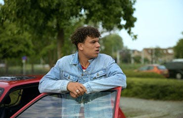 Een jonge man in een spijkerjack staat naast zijn auto en leunt met zijn armen op het openstaande portier. 