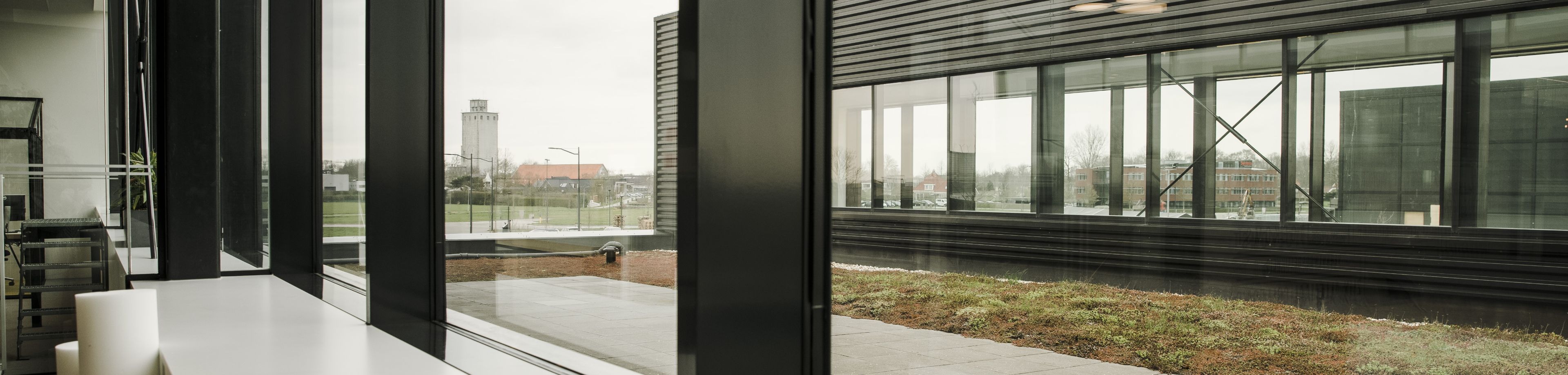 Een foto die door het raam van een bedrijfspand genomen is. Van binnen naar buiten kijkend, op de foto zie je een groen dak en een ander deel van het bedrijfspand.