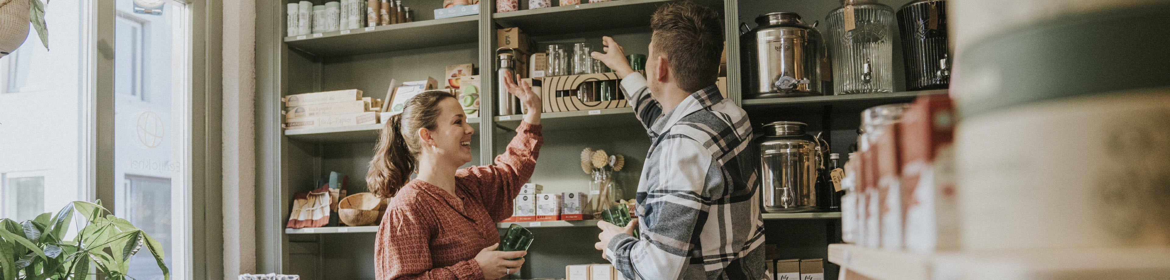 Een verkoopmedewerker en een klant in een sfeervolle winkel reiken beiden met hun hand naar een plank met producten.