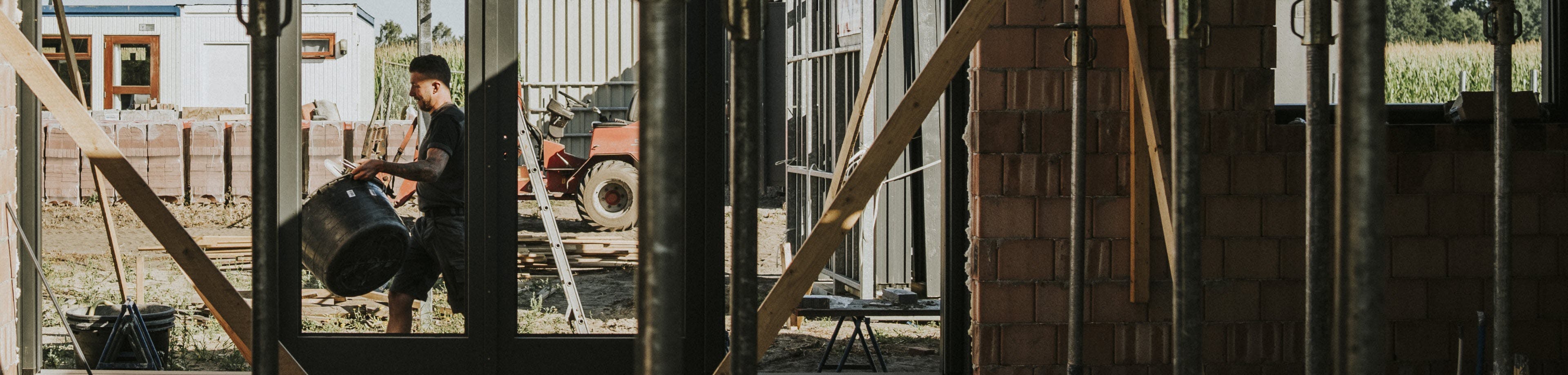 Een man in werkkleding loopt met een speciekuip over een bouwplaats. De foto is gemaakt van binnenuit een pand in aanbouw.
