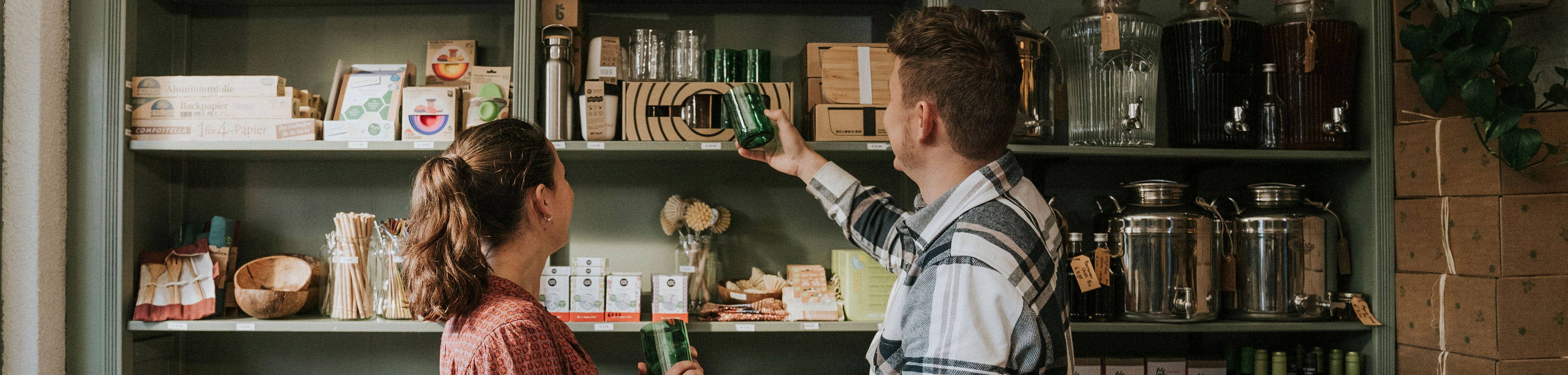 Een man en een vrouw staan bij een winkelkast vol spullen, en bekijken een metaalkleurig item uit die kast.