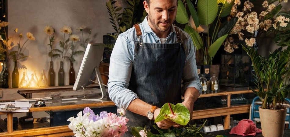Een man staat met een schort aan in een bloemenzaak en werkt aan het samenstellen van een boeket.