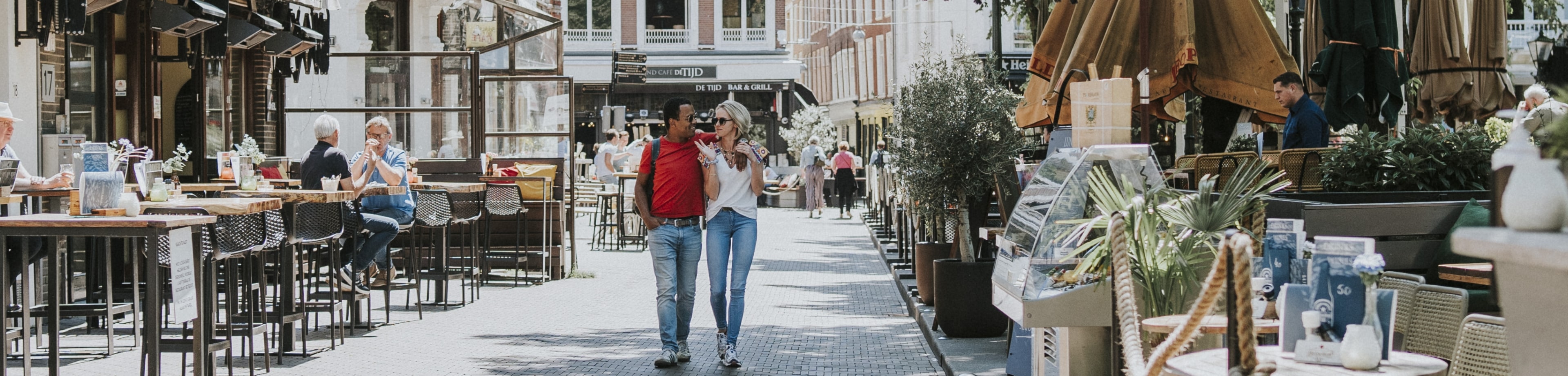 Een man loopt vrolijk met zijn arm om de schouder van een vrouw over straat, langs terrasjes. De zon schijnt. 