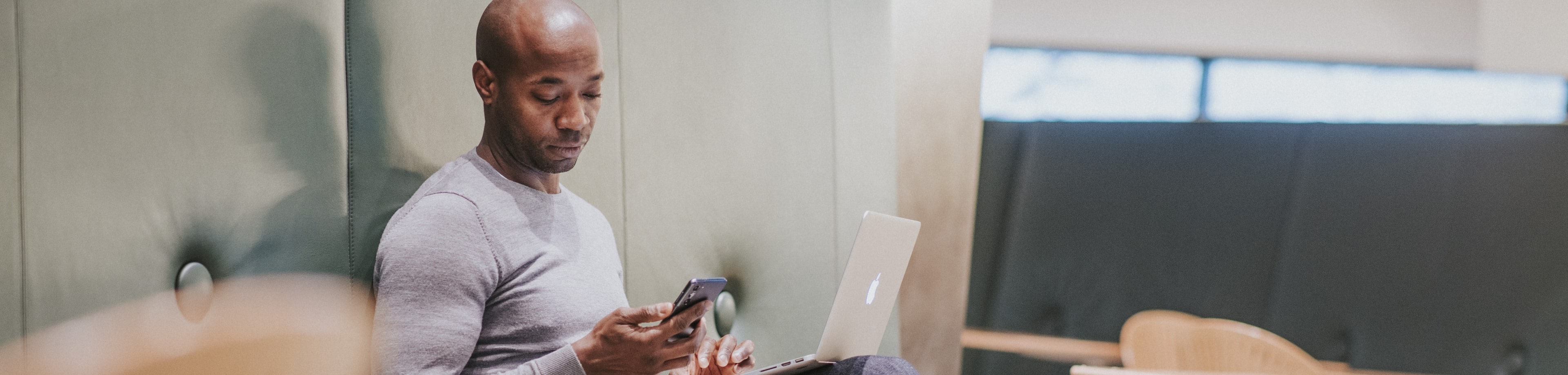 Een man zit op een groene bank, met zijn laptop op schoot en zijn smartphone in de hand. Hij kijkt op het scherm van zijn telefoon.
