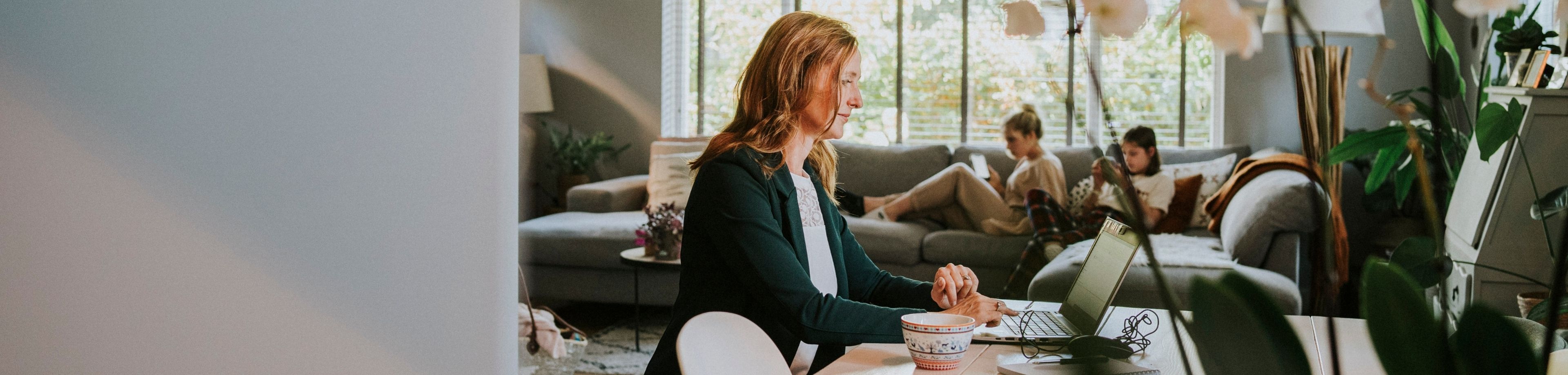 Vrouw werkt aan eetkamertafel en dochters zitten op de bank