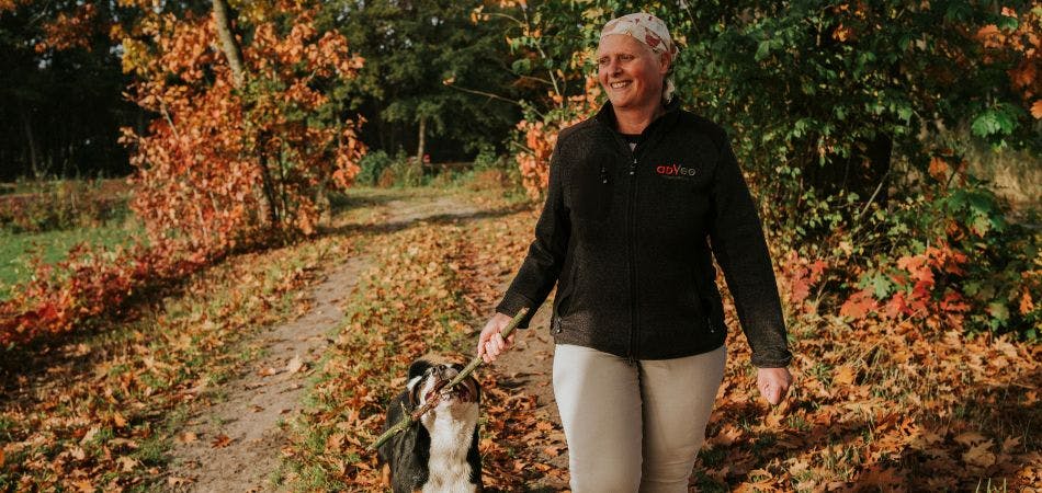 Dierenarts Janny Hermans loopt met haar hond door een bosrijke omgeving. Ze lacht ontspannen en houdt een stok in haar hand; de hond heeft diezelfde stok tegelijkertijd in zijn bek.  