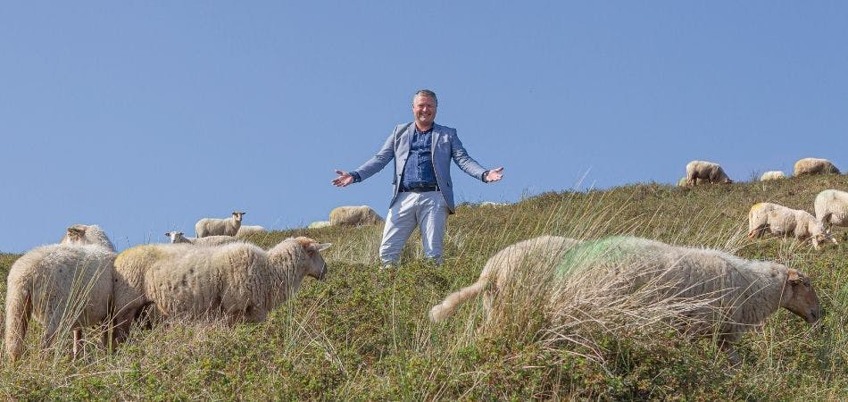 Vrolijke John, met zijn armen gespreid en lachend tussen de schapen op een dijk
