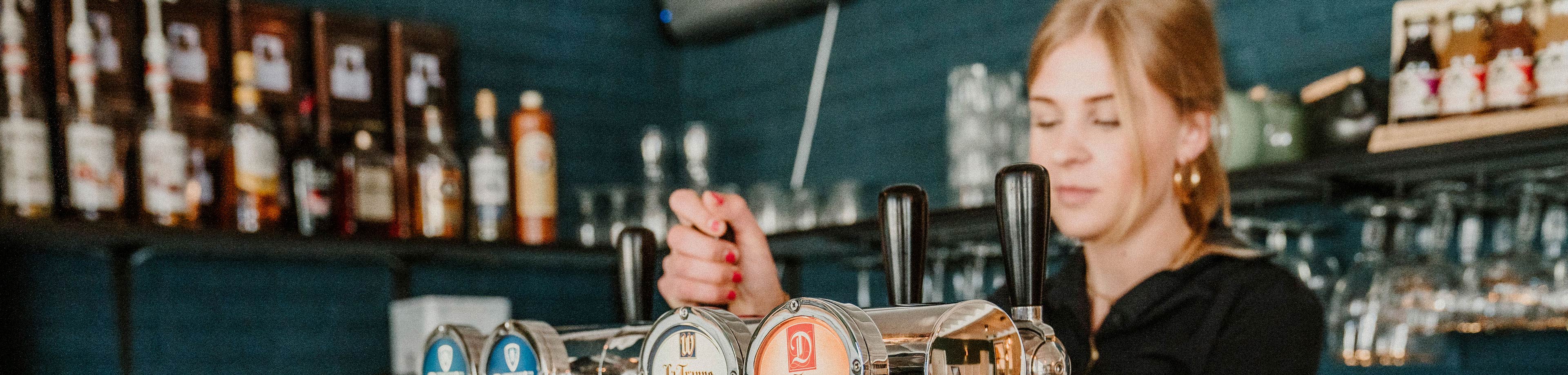 Een jonge vrouw tapt een biertje achter de bar van een horeca-gelegenheid.