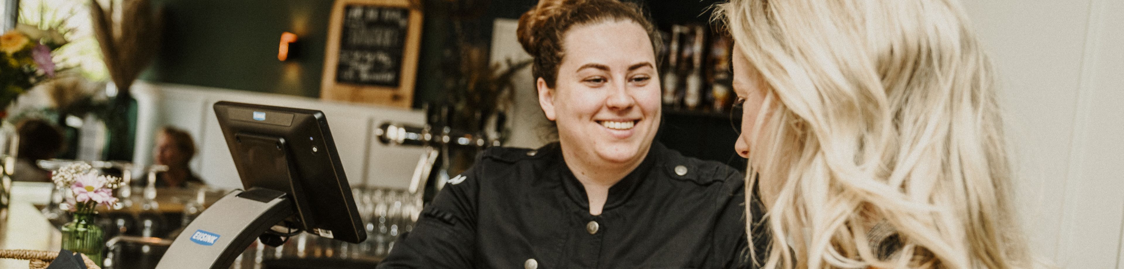 Een klant van een horeca-gelegenheid rekent met haar pinpas af bij een vrolijk lachende medewerker.