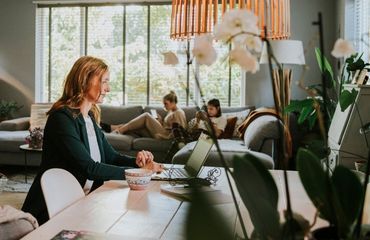 Een vrouw werkt aan een eetkamertafel op haar laptop. Op de achtergrond zitten haar dochters op de bank.