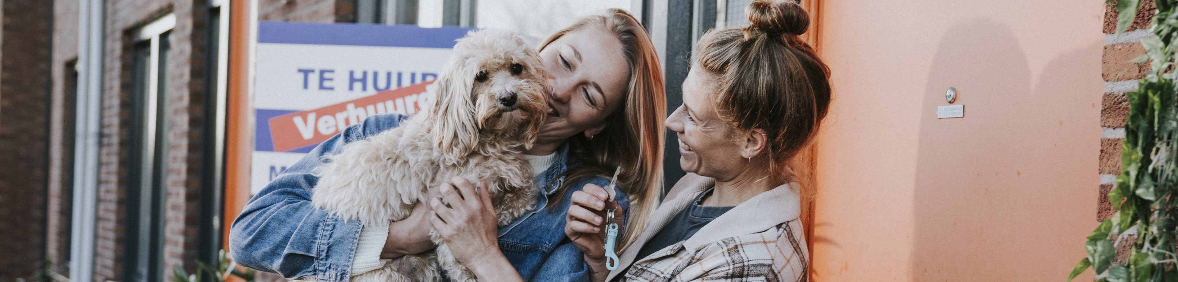Een jong stel staat met hun hondje voor de deur van hun nieuwe huurwoning.