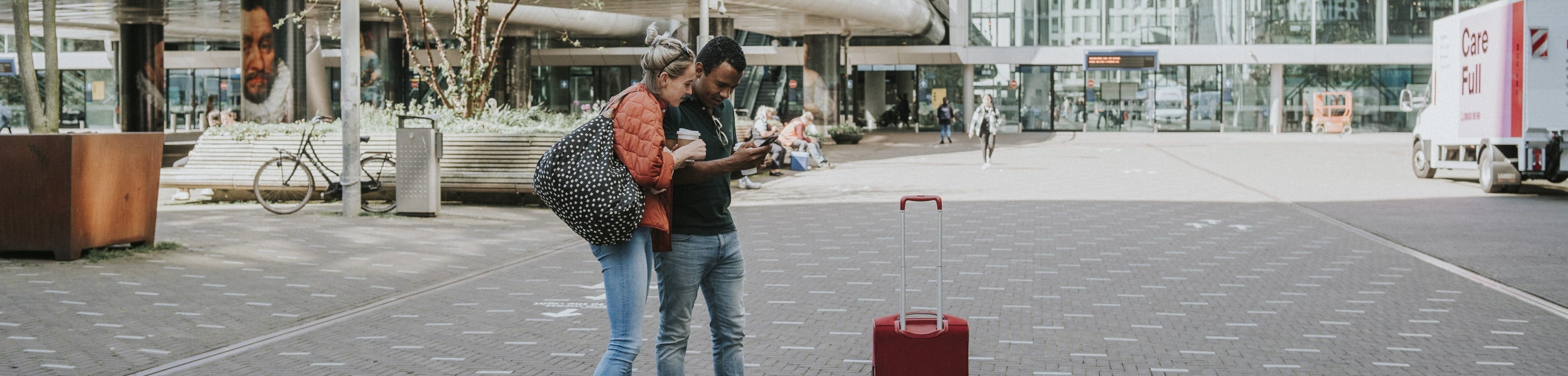 Man en vrouw staan met koffie in de hand en bagage op een stationsplein en kijken op de telefoon die de man vasthoudt