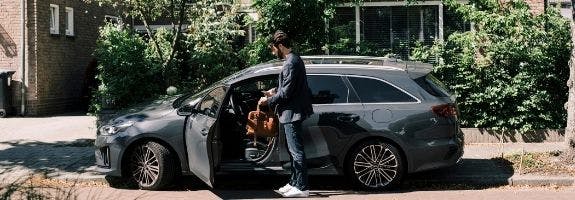 Een man staat naast zijn auto met geopend portier. Hij heeft zijn tas en telefoon in de hand, en is klaar om in te stappen.