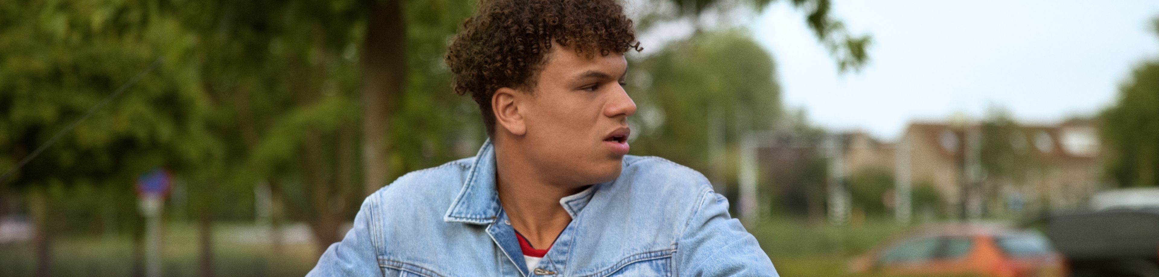 Een jonge man in een spijkerjack hangt met zijn armen over een openstaand autoportier.