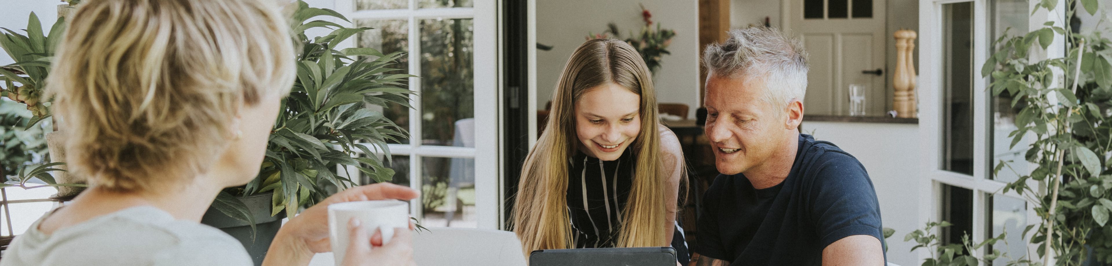 Vader zit samen met dochter met een laptop aan tafel. Moeder zit erbij met een kop thee. 