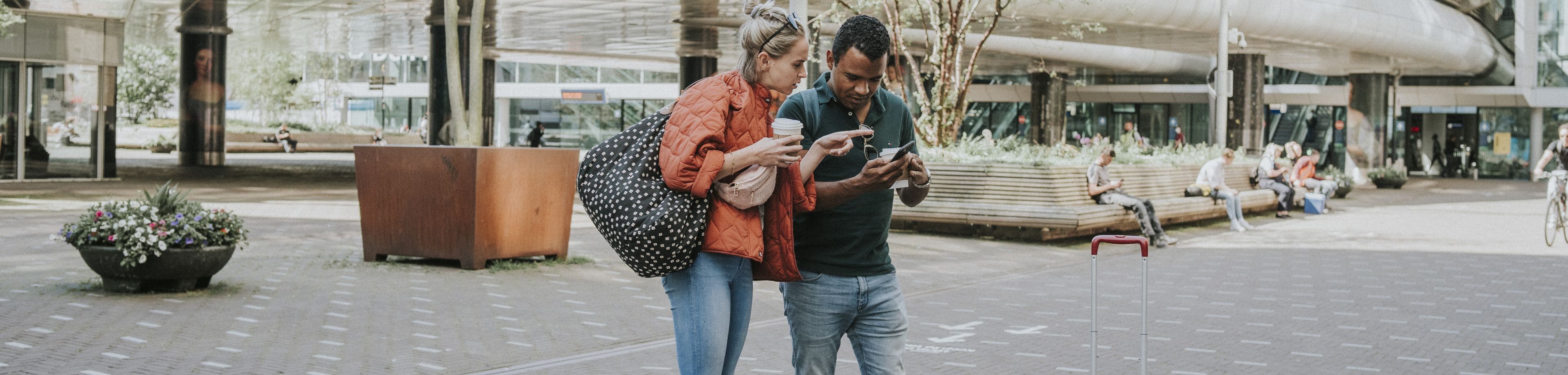 Een stel staat op een stationsplein aan het begin van een reis. Ze kijken samen naar het scherm van een mobiele telefoon ter voorbereiding op hun reis.