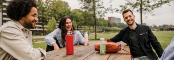 Een groepje jonge mensen zit bij elkaar aan een houten picknicktafel in een stadspark.