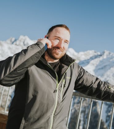 Een man staat te bellen bij een hek met besneeuwde bergen op de achtergrond.