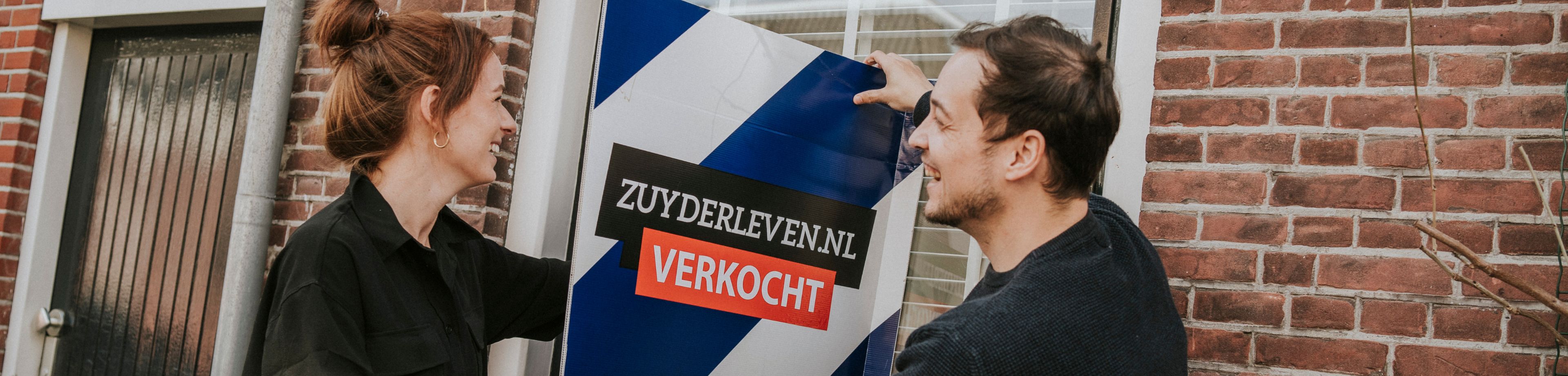 Man en vrouw lachen en houden een verkocht bord vast bij het raam van een woning