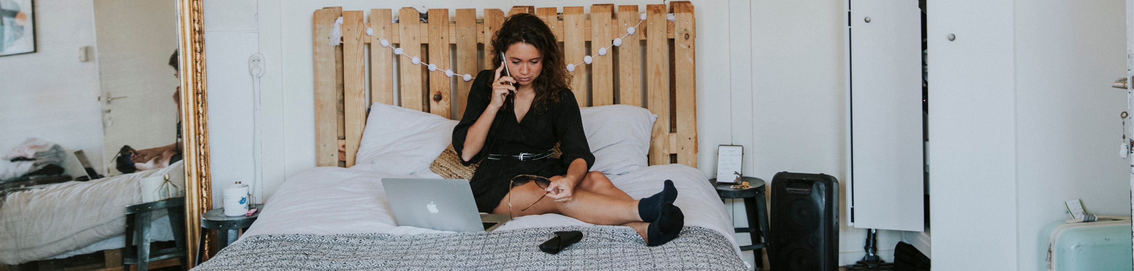 Vrouw zit op bed te bellen met in haar hand een zonnebril, terwijl op het bed een opengeklapte laptop staat.