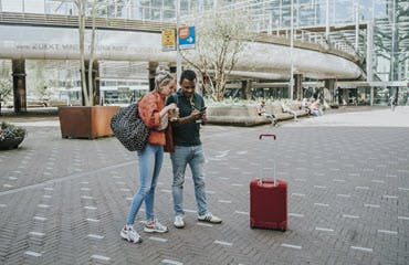 Twee mensen staan met een koffer voor een stationsgebouw, en zoeken iets op op een mobiele telefoon.