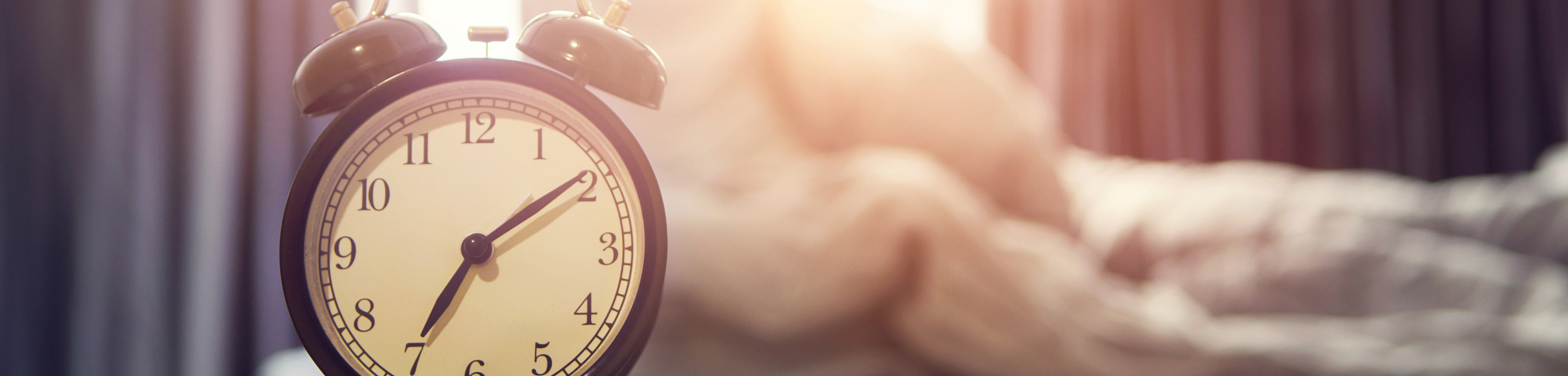 Wekker met op de achtergrond een vrouw die zich uitrekt terwijl ze op bed zit.