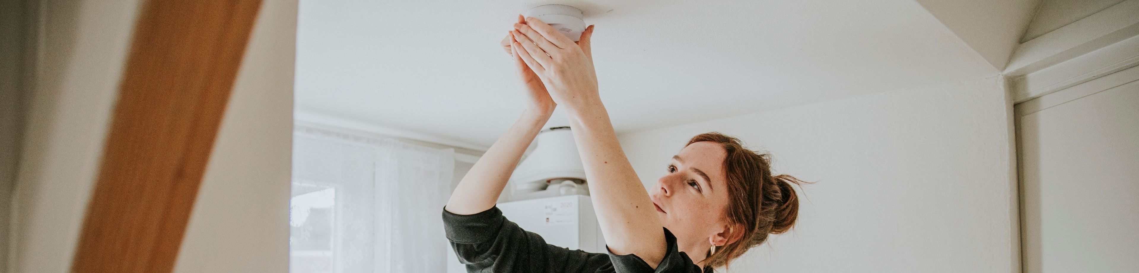 Een vrouw controleert of bevestigt een rookmelder aan het plafond. Het is belangrijk om regelmatig je rookmelders te testen, schoon te maken en te onderhouden om zeker te weten dat deze je op tijd waarschuwen in geval van rook of brand