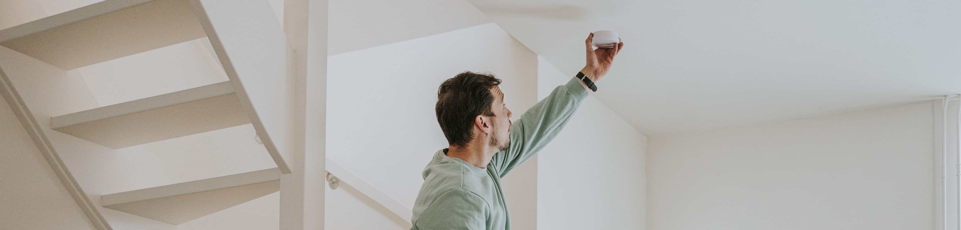 Man staat op trap en houdt een rookmelder tegen het plafond en vrouw kijkt naar de rookmelder