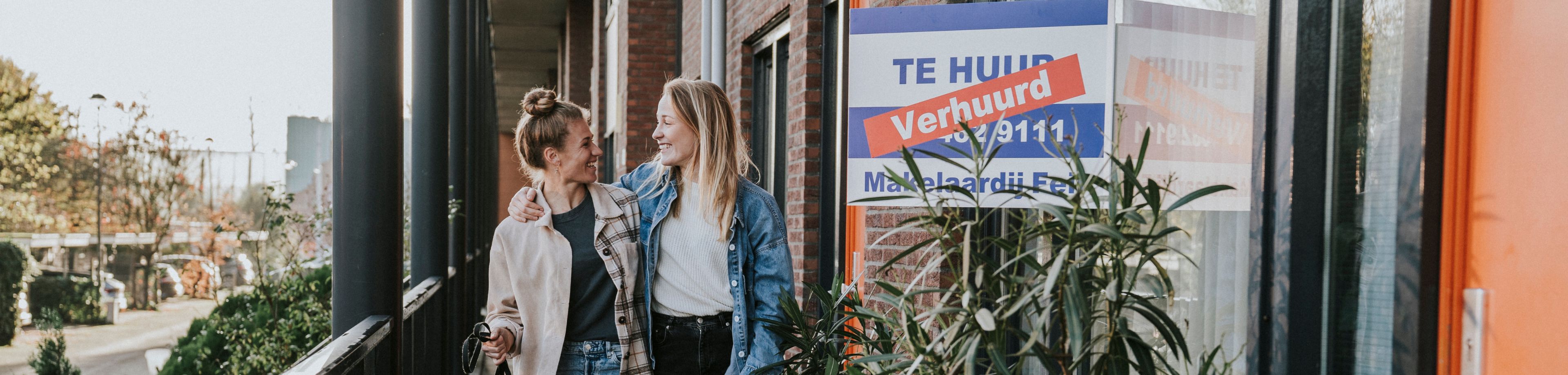 2 vrouwen lopen met een aangelijnde hond over de galerij van een appartementencomplex. Ze kijken elkaar aan en hebben een lach op hun gezicht. Op het appartement waar ze langslopen, hangt een bord met 'Verhuurd' aan het raam.