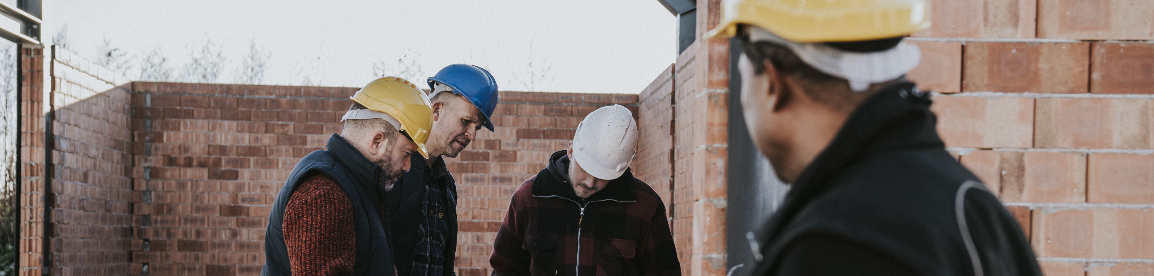 Vier mensen staan op de bouwplaats en kijken samen naar bouwtekeningen.
