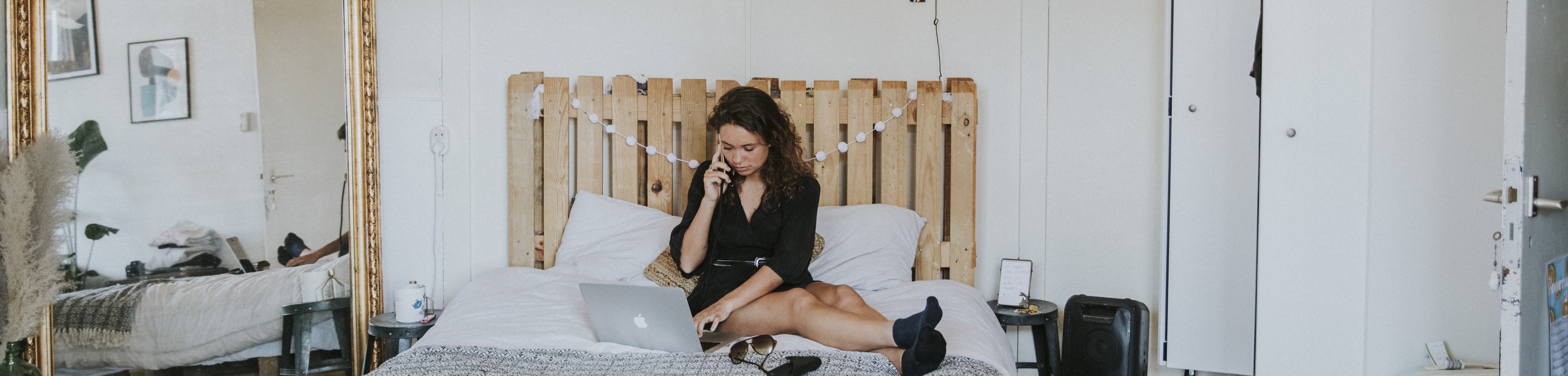 Een vrouw zit met de laptop opengeklapt op het bed, ze heeft een telefoon aan haar oor.