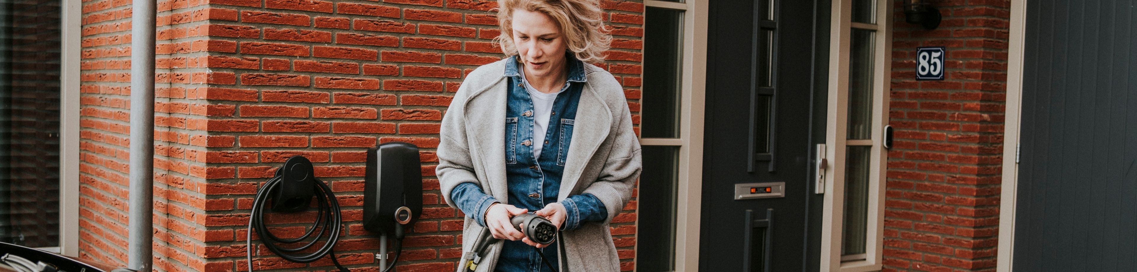 Een vrouw sluit haar elektrische auto aan op de laadpaal, die aan de gevel van het woonhuis hangt.