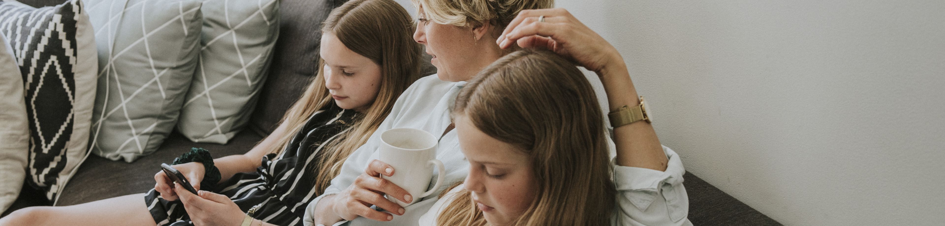 Een vrouw zit op de bank tussen haar beide dochters in. Ze heeft een witte mok vast, en de dochters kijken op hun mobiele telefoons.