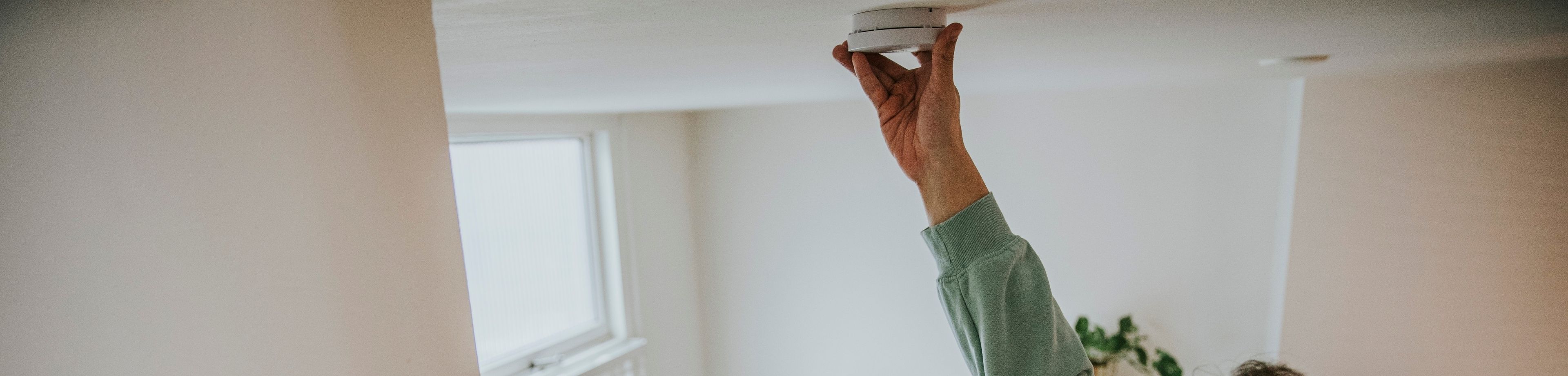 Een man staat op een trapje en hangt een rookmelder op aan het plafond.