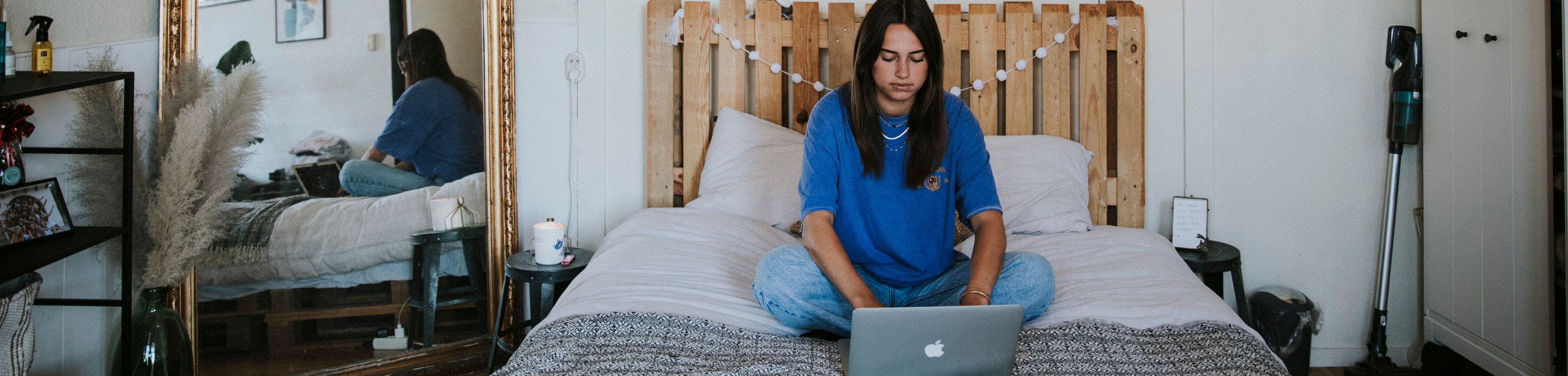 Een jonge vrouw zit op haar bed. Ze is online aan het shoppen op haar laptop.