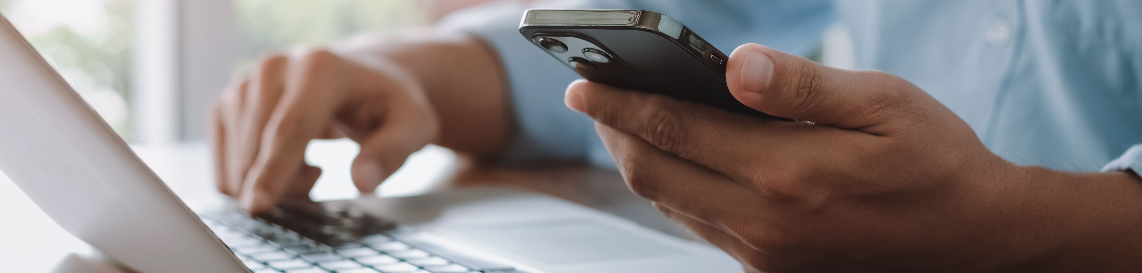 Foto van een persoon die achter een laptop werkt. De foto is afgesneden zodat je alleen de handen ziet. Deze persoon heeft in de linkerhand een telefoon.