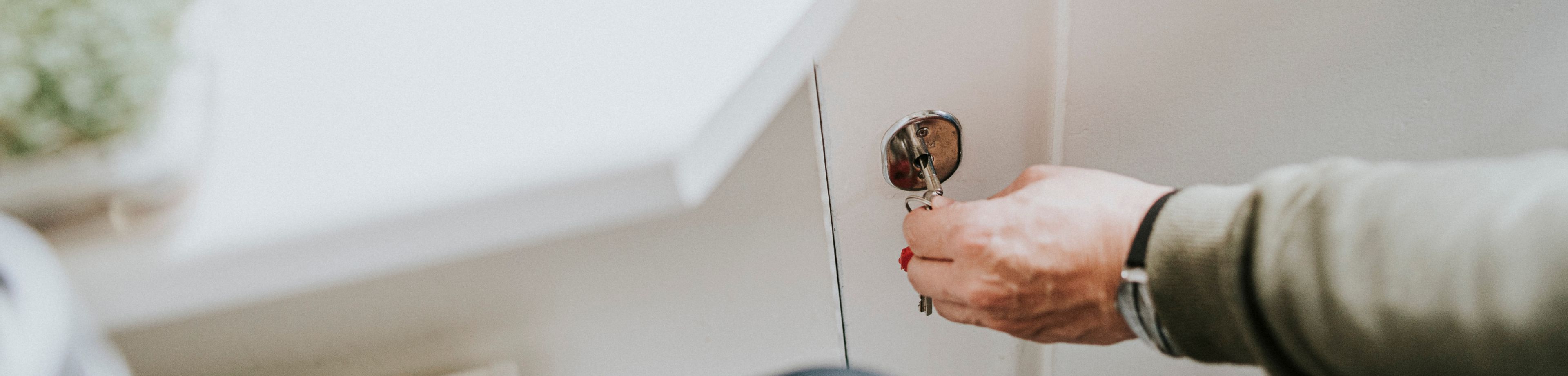Een persoon houdt met 1 hand de klink van een witte buitendeur vast, en draait met de andere hand de sleutel om in het slot.