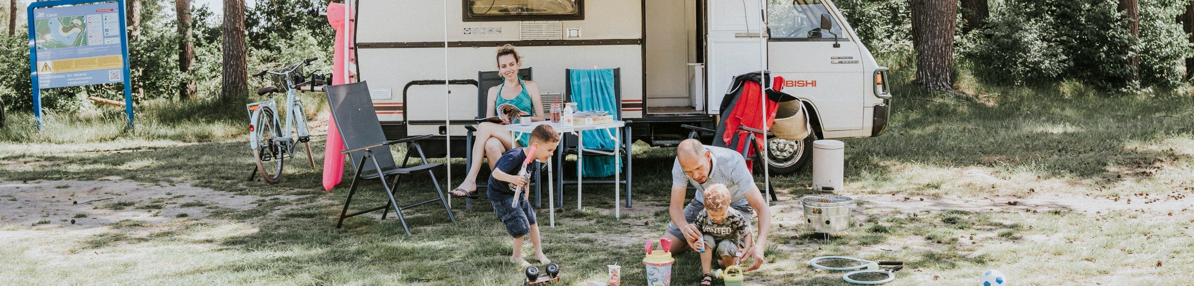 Gezin is aan het kamperen, vrouw zit een tijdschrift te lezen, man en kinderen spelen voor de camper
